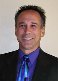 Dr. greg Malakoff with short, dark hair wearing a black suit jacket, a purple dress shirt, and a colorful blue and purple tie, smiling at the camera against a plain light background.