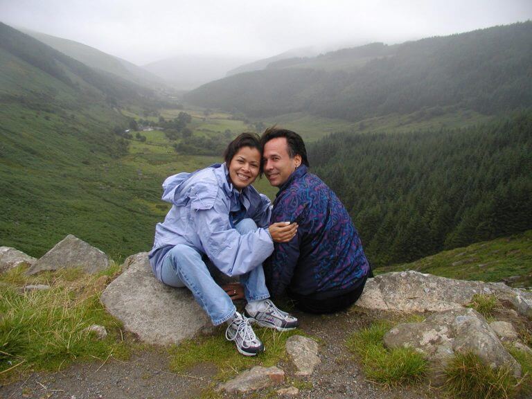 Dr. Malakoff and his wife Pam - smiling as the sit on rocks overlooking a lush, green valley with rolling hills and forests under a cloudy sky. The woman, dressed casually in a light jacket and sneakers, relaxes beside her partner.
