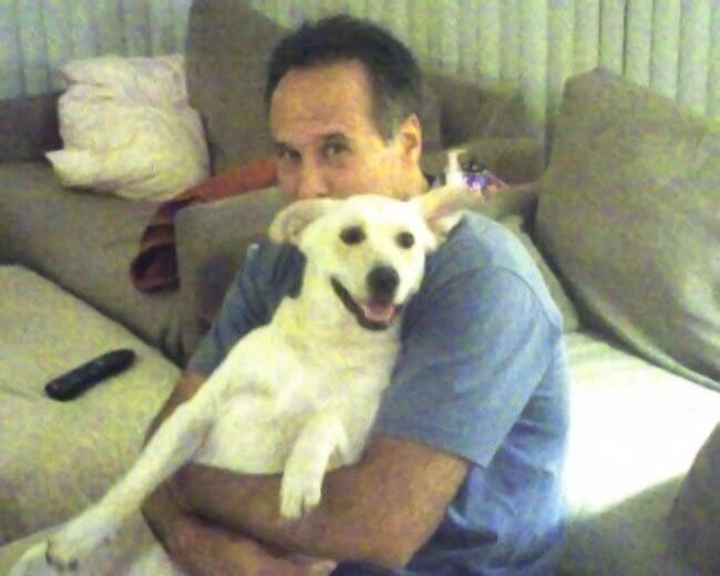 Dr. Greg Malakoff, Chiropractor sits on a couch hugging a happy, white dog with large ears. The man is wearing a blue shirt, and there is a remote control and pillows on the couch beside them.