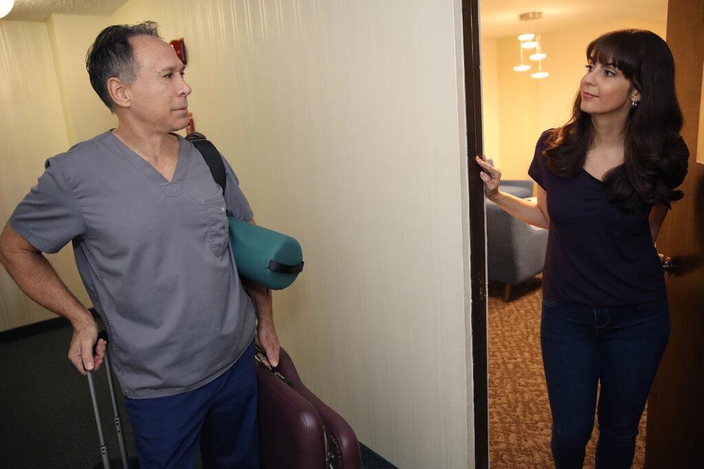 Dr. Greg Malakoff, Chiropractor, in scrubs, possibly a chiropractor or dr, carrying a suitcase and gym bag stands in a hallway, looking at a woman with long dark hair who is smiling and standing in the doorway of her Los Angeles apartment.