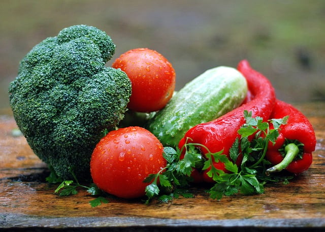 A group of fresh vegetables on a wooden surface, including broccoli, two red tomatoes, a cucumber, two red bell peppers, and sprigs of parsley with drops of water.