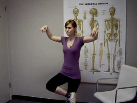 A woman stands on one leg in an office, with arms bent and pointed downward, mimicking a bird pose. She is in front of a skeletal system poster at a Los Angeles chiropractor’s clinic, near a white chair.