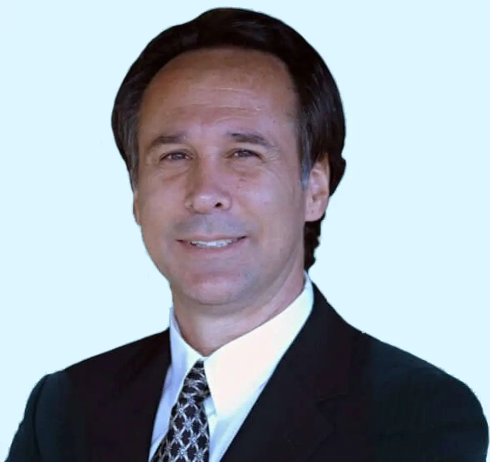 Dr. Greg Malakoff with dark hair wearing a dark suit, white shirt, and patterned tie, smiling slightly against a light blue background, presenting the professional demeanor of a doctor.