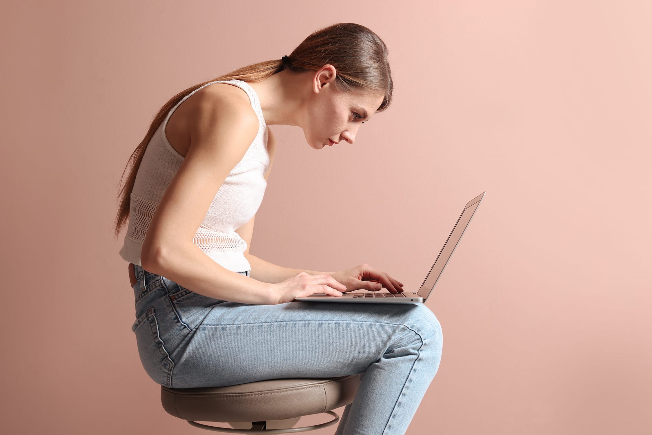 A young woman with long hair sits on a stool, hunched forward with poor posture while typing on a laptop. She wears a white sleeveless top and light blue jeans, against a plain peach background.