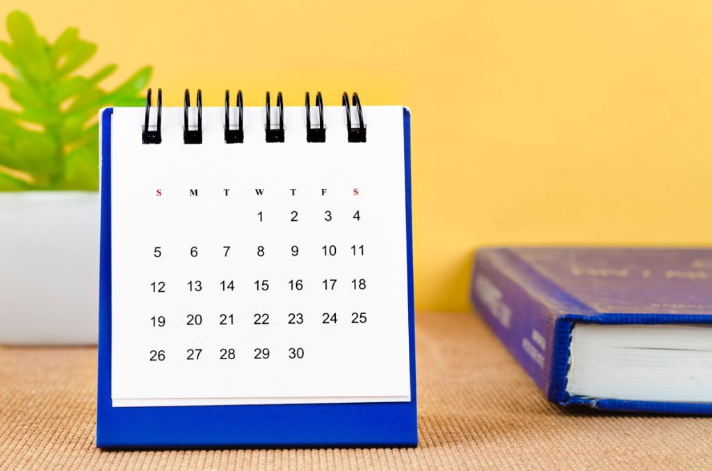 A small desk calendar with a blue stand shows a month, sitting on a table next to a hardcover book at a Los Angeles chiropractor’s office, alongside a green potted plant and a yellow background.